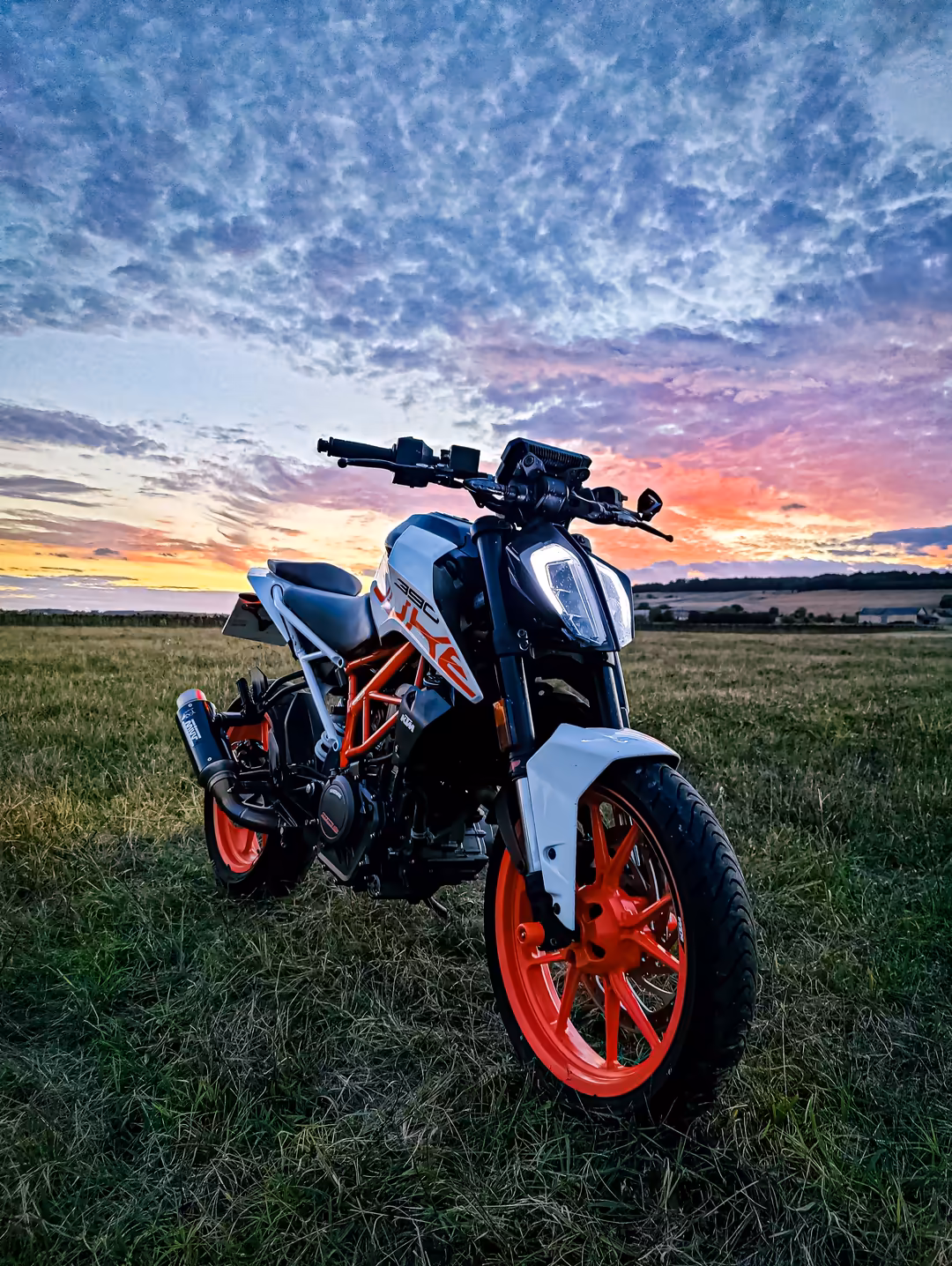 Red and white motorcycle parked in a grassy field under a dramatic sunset sky with colorful clouds