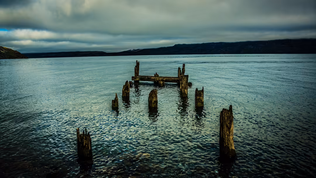 Weathered wooden pilings jutting from calm turquoise water beneath a moody cloudy sky, with distant shoreline visible