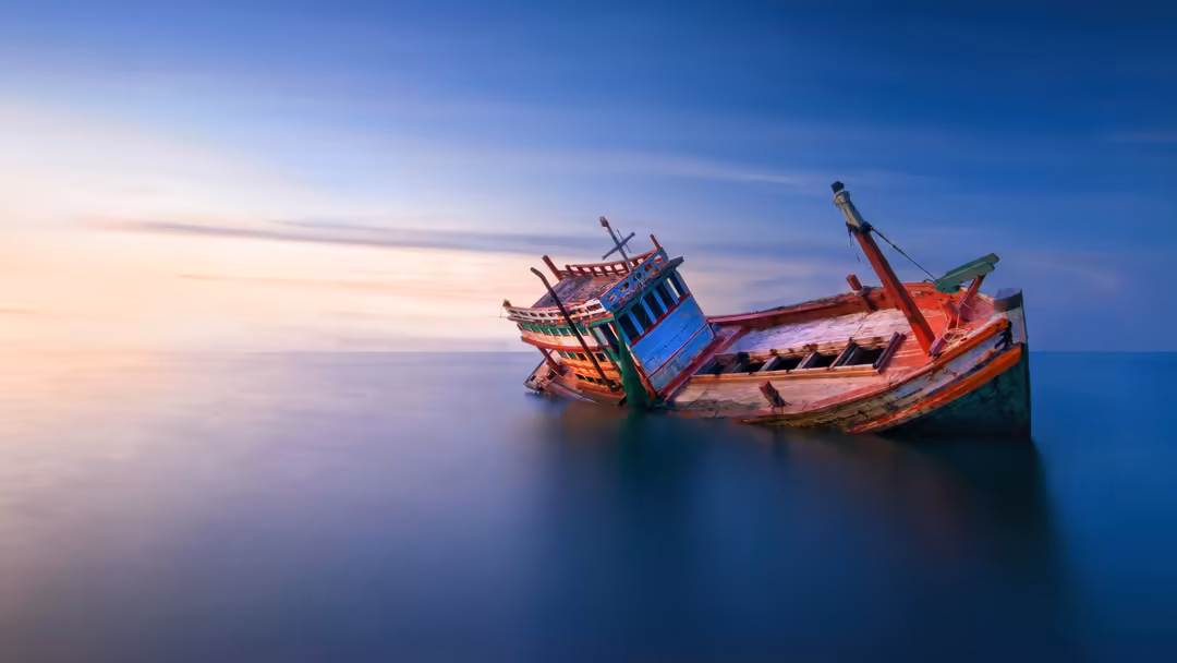 Abandoned, colorful wooden fishing boat sinking in calm blue ocean waters at sunset with serene sky reflecting on still water