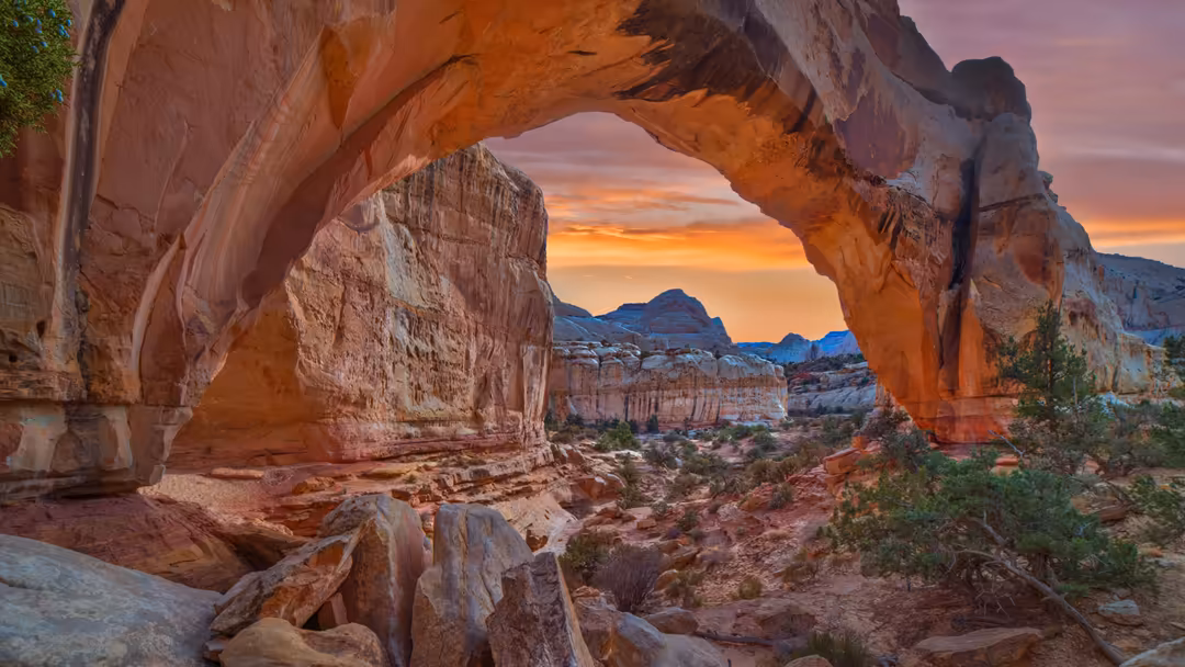Natural stone arch framing a dramatic sunset over desert landscape with red rock formations and distant mountains