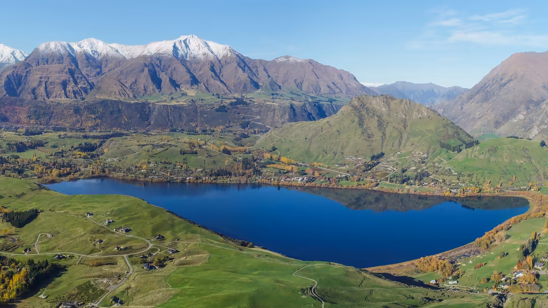 Aerial view of a pristine alpine lake nestled in a verdant valley, surrounded by snow-capped mountains and scattered farmhous