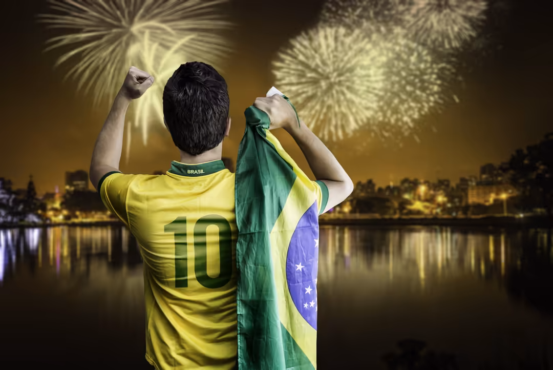 Soccer player in yellow Brasil jersey holding national flag with arms raised, celebrating with golden fireworks exploding ove