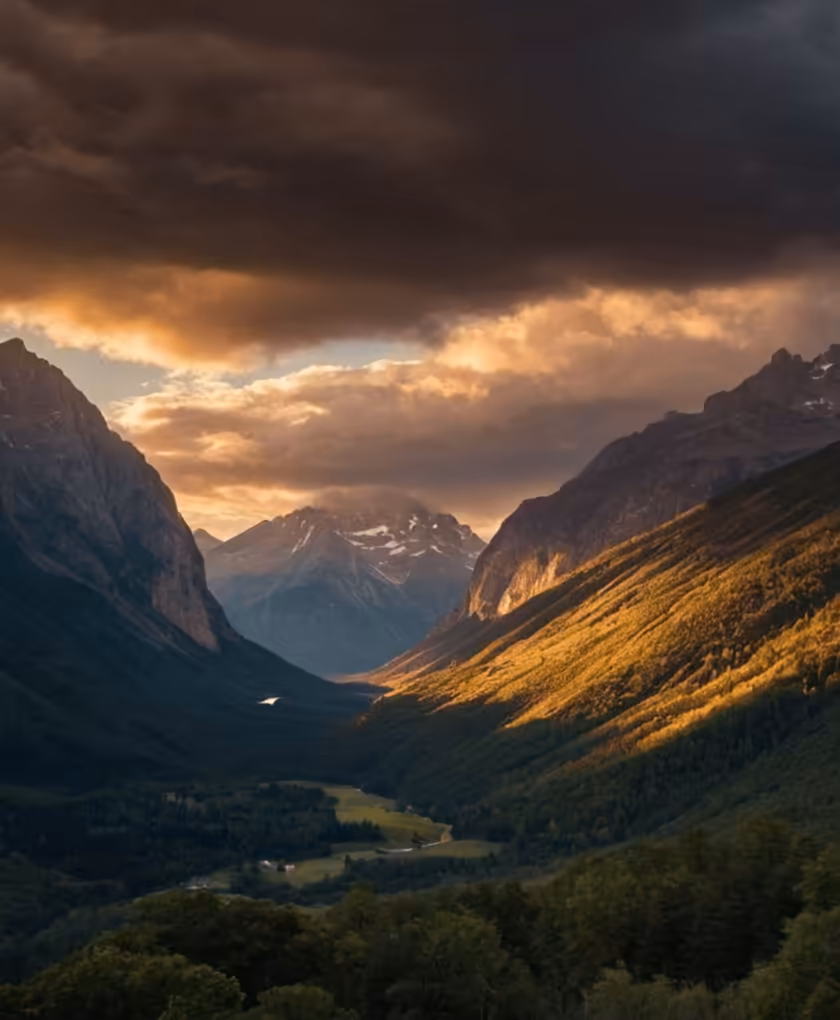 Dramatic alpine valley with towering cliff mountains and golden-hour sunlight illuminating forested slopes, snow-capped peaks