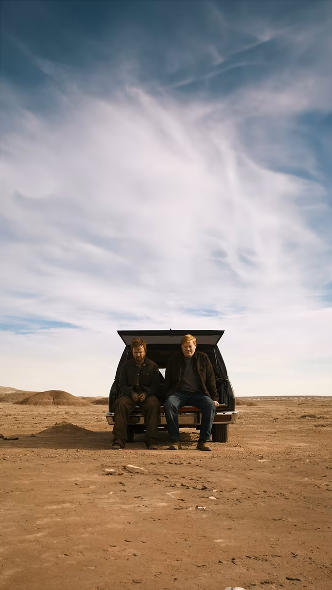 Two men sit in the open trunk of a car on a vast desert landscape under a dramatic cloudy sky, evoking a tense moment from th