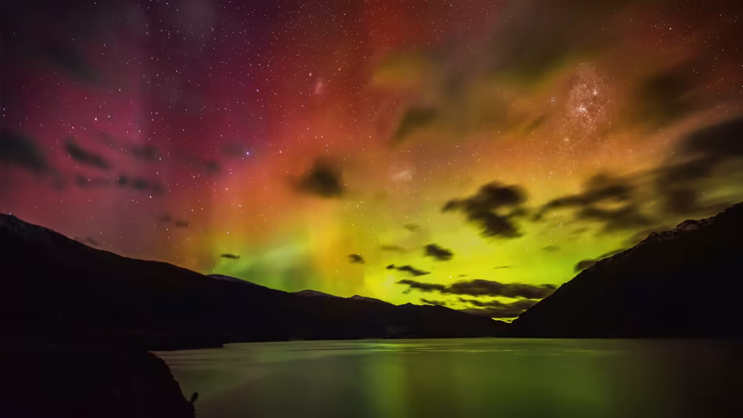 Vibrant aurora borealis with green and pink lights dancing across the night sky above a serene mountain lake with silhouetted