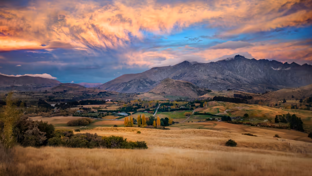 Dramatic sunset sky with golden clouds above a verdant valley dotted with trees, winding river, and rugged mountain peaks in