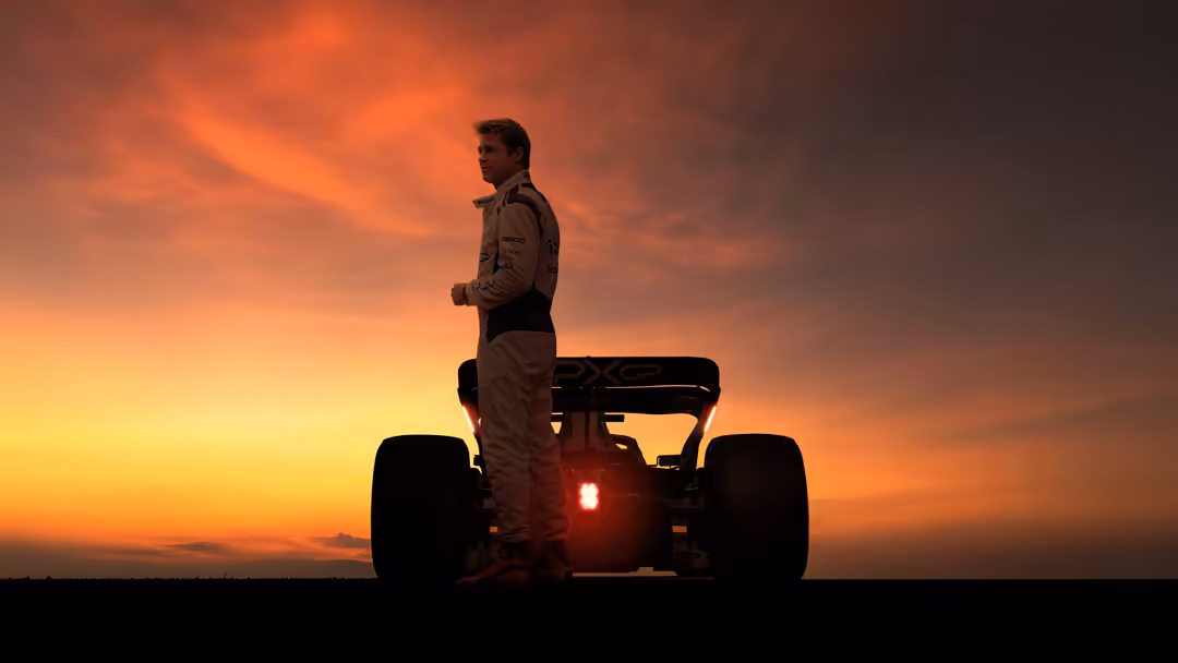Formula 1 driver standing on racing car silhouetted against dramatic orange and yellow sunset sky with glowing tail light