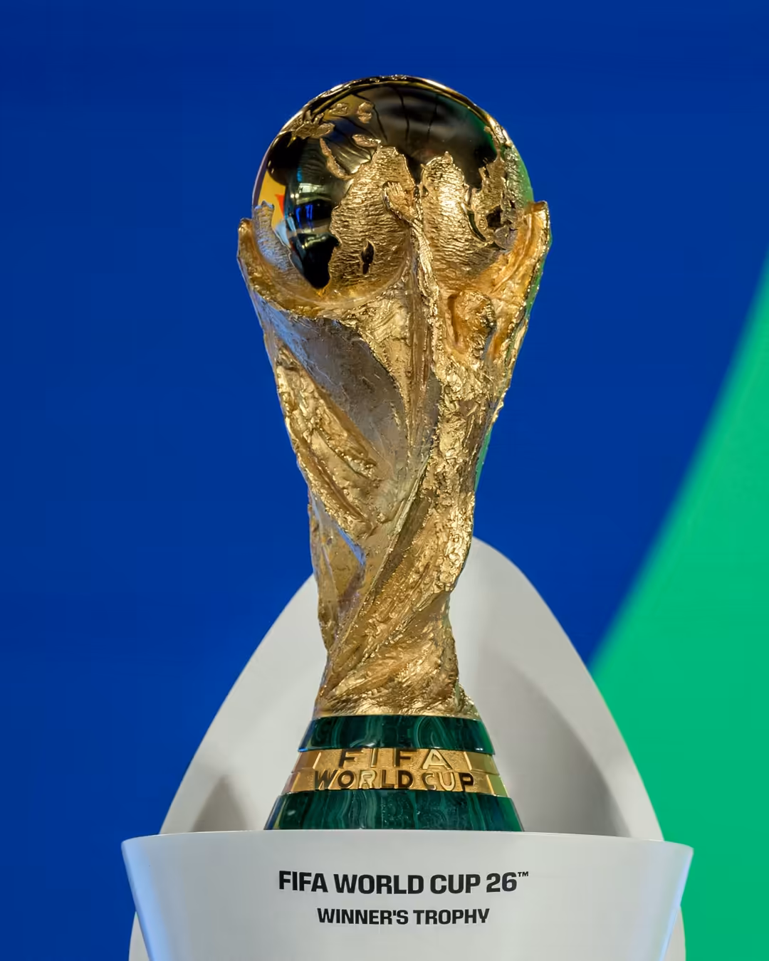Golden FIFA World Cup 26 winner's trophy displayed against a vibrant blue and green background, featuring the iconic statue a