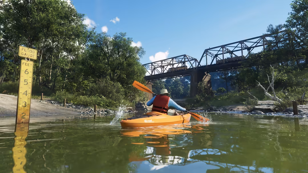 Player kayaking in orange kayak on green river with industrial bridge and lush trees under blue sky