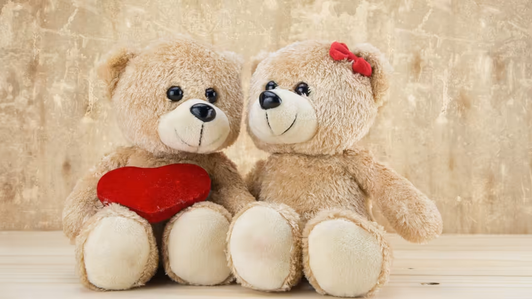 Two adorable beige teddy bears sitting together, one holding a red heart, wearing a red bow, on textured beige background