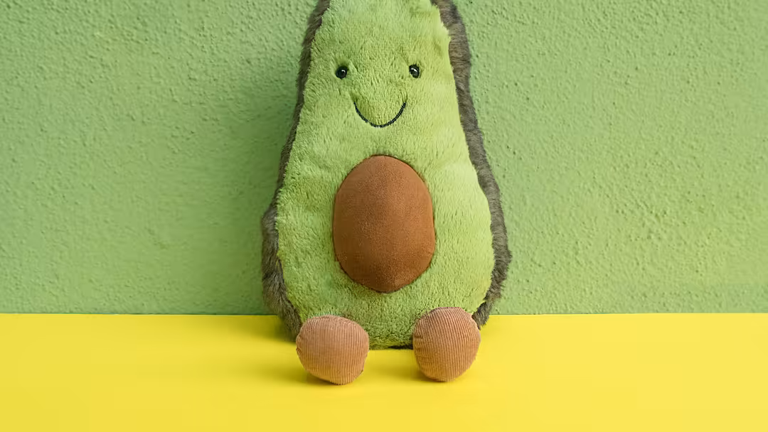 Adorable green plush avocado toy with smiling face and brown pit sitting on yellow surface against green wall