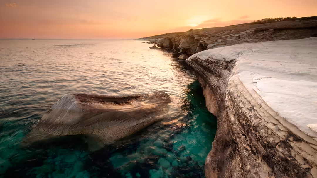 Rocky coastal cliffs at golden hour sunset with turquoise water, white limestone formations, and warm peach-colored sky refle