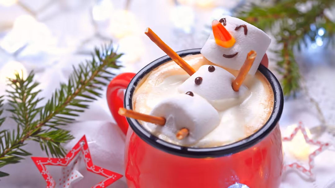 Adorable marshmallow snowman in a red mug of hot chocolate with cinnamon stick, evergreen sprigs, and festive red star decora