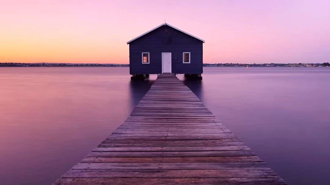 A serene boathouse with a wooden dock stretching across calm purple water at sunset, surrounded by a pink and golden sky.