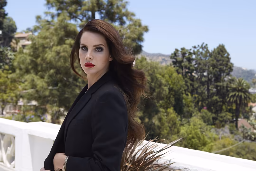 Woman with long brunette waves and red lipstick wearing black blazer, posed against lush green trees and hillside landscape i