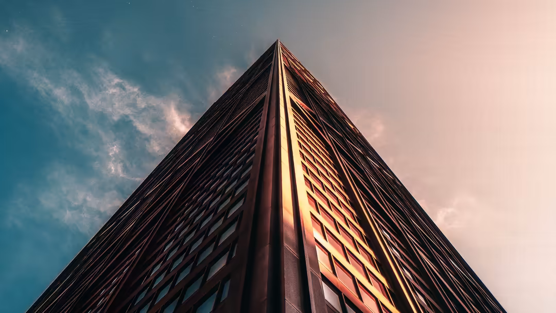 Towering modern building shot from below against blue sky with golden sunlight illuminating its geometric facade and sharp ed