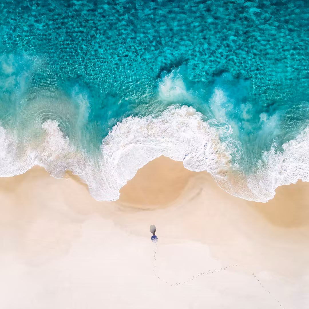 Aerial view of a person standing on pristine sandy beach with turquoise ocean waves and white foam crashing on shore