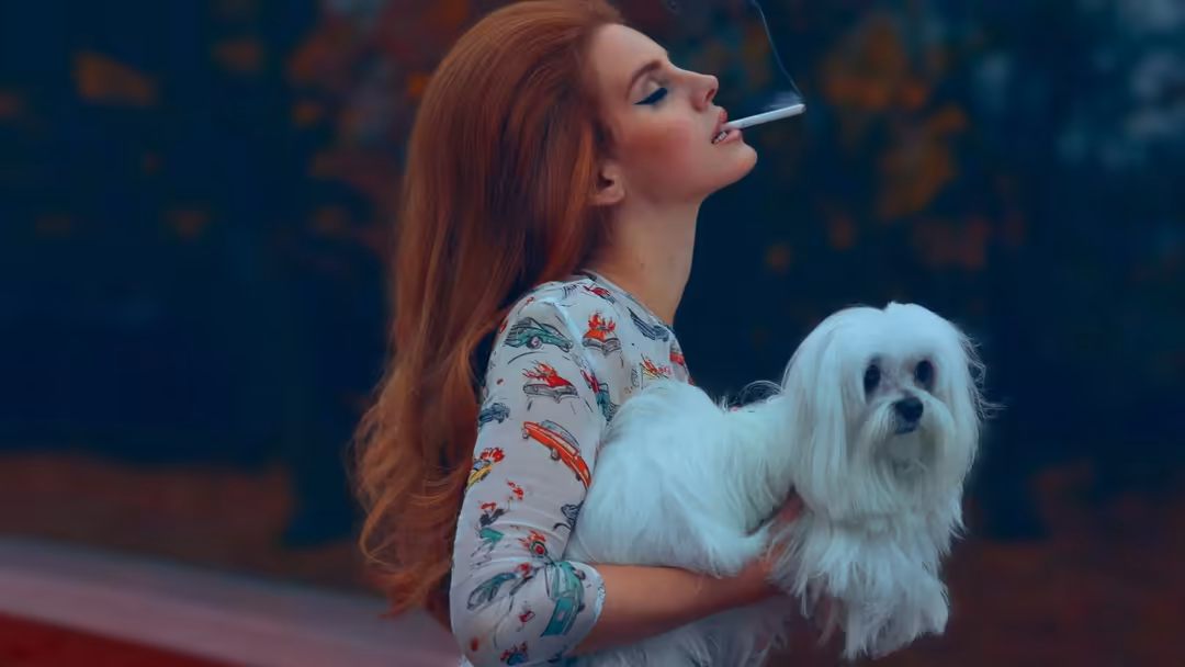 Woman with long red hair holding a white fluffy dog, smoking cigarette, wearing patterned shirt against dark background with