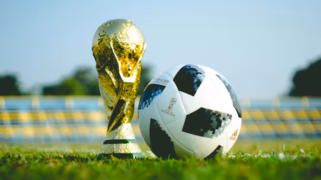 Golden FIFA World Cup trophy and classic black-and-white soccer ball displayed on green grass field with stadium background