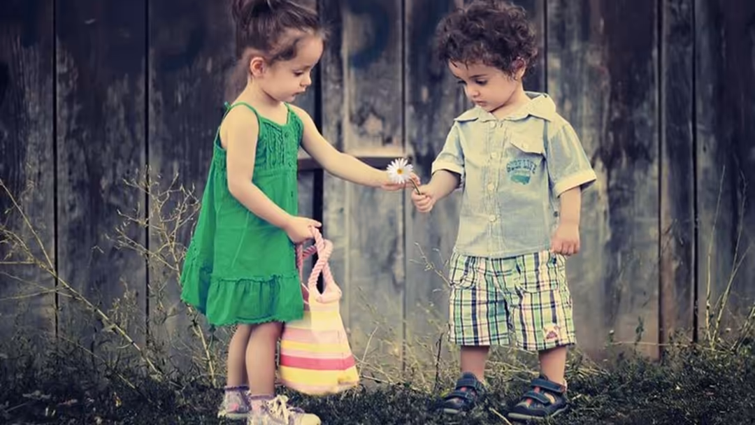Two young children in vintage clothing sharing a small daisy flower against a weathered wooden backdrop, capturing a sweet mo