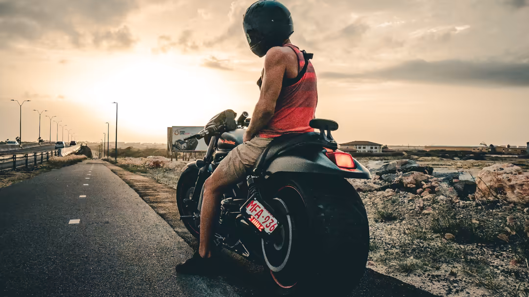 Motorcyclist in red shirt riding on desert highway at sunset with street lights and distant buildings along the road