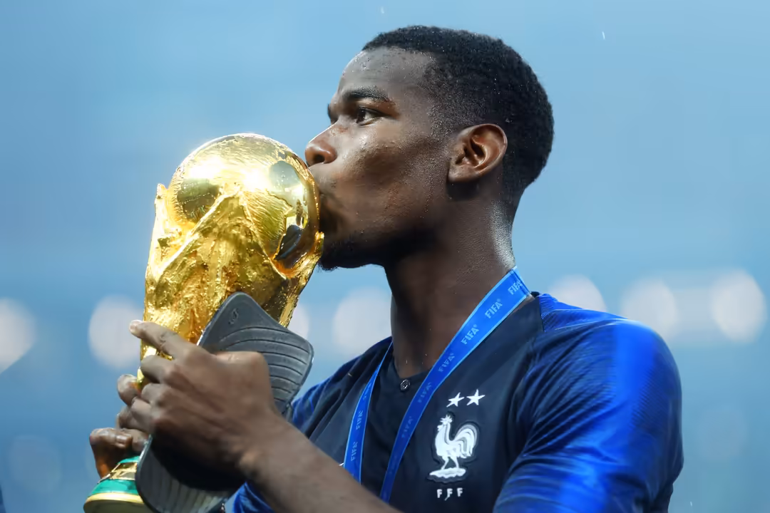 Athlete in blue jersey holding golden FIFA World Cup trophy against clear sky with medal around neck
