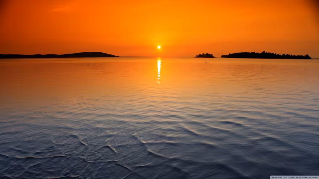Golden sunset reflecting across calm ocean waters with forested islands silhouetted against an orange and yellow sky