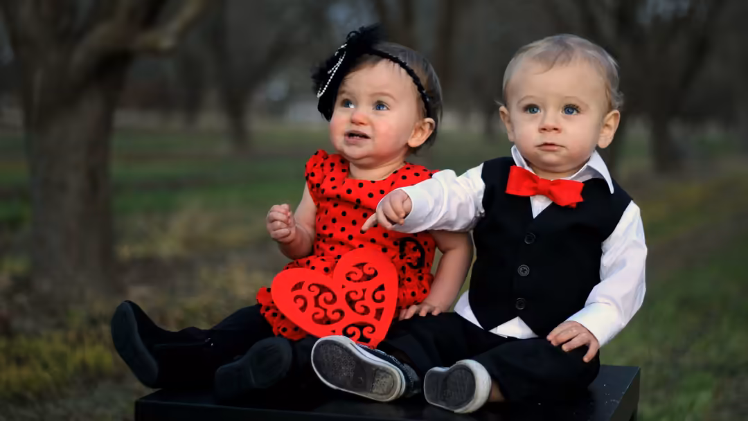 Two dressed toddlers sitting together outdoors, one in red polka dot dress with bow, one in formal vest and red bow tie