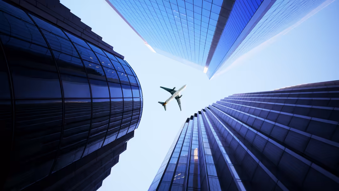 Commercial airplane flying overhead between tall modern glass skyscrapers against clear blue sky in urban cityscape