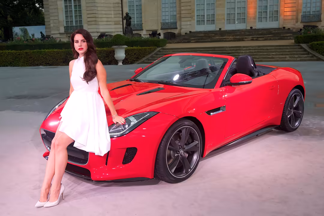 Woman in white dress posing beside a red convertible sports car in front of a classical building with manicured hedges