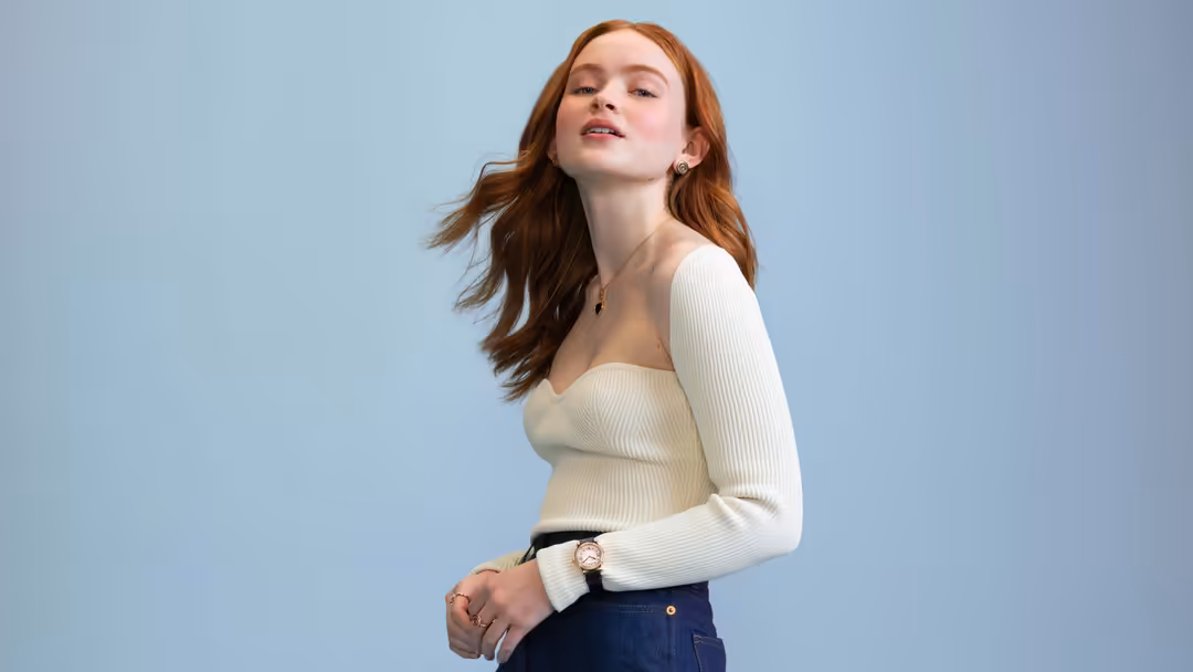 Redheaded woman in cream ribbed sweater and dark jeans against light blue background, wearing watch and small necklace, looki