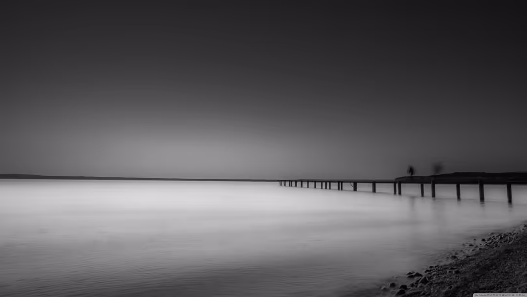 Minimalist black and white long exposure of wooden pier extending into calm misty water with silhouetted figures under overca