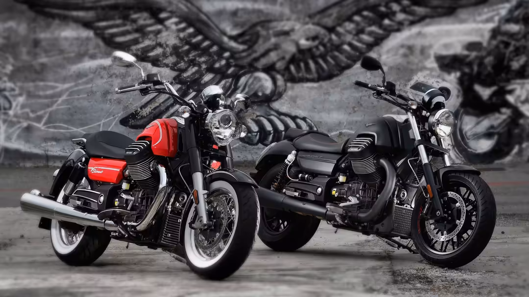Two classic cruiser motorcycles parked side by side, one red and black, one black, with chrome details and white-wall tires a