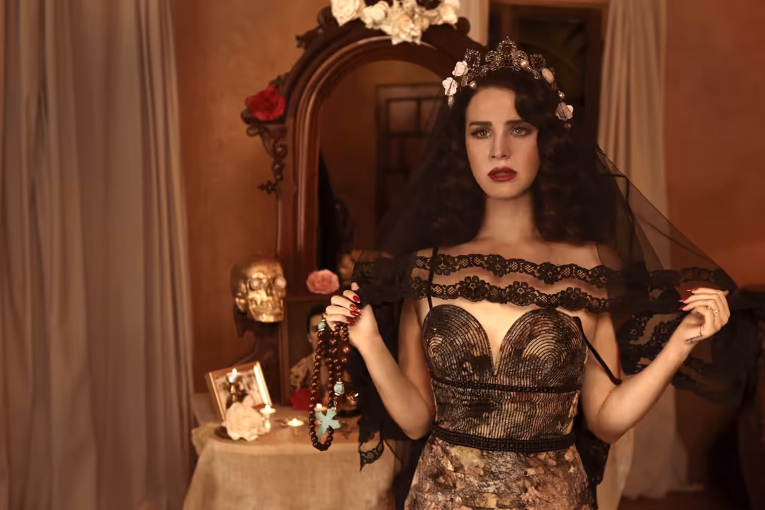 Woman in black lace off-shoulder dress with flower crown and veil, holding beaded necklace in ornate vintage room with warm l