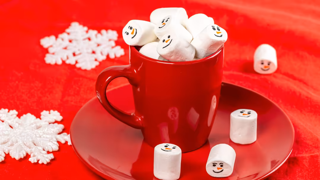 Cheerful marshmallows with smiling faces in a red mug on a red saucer, surrounded by white snowflakes and snowman decorations
