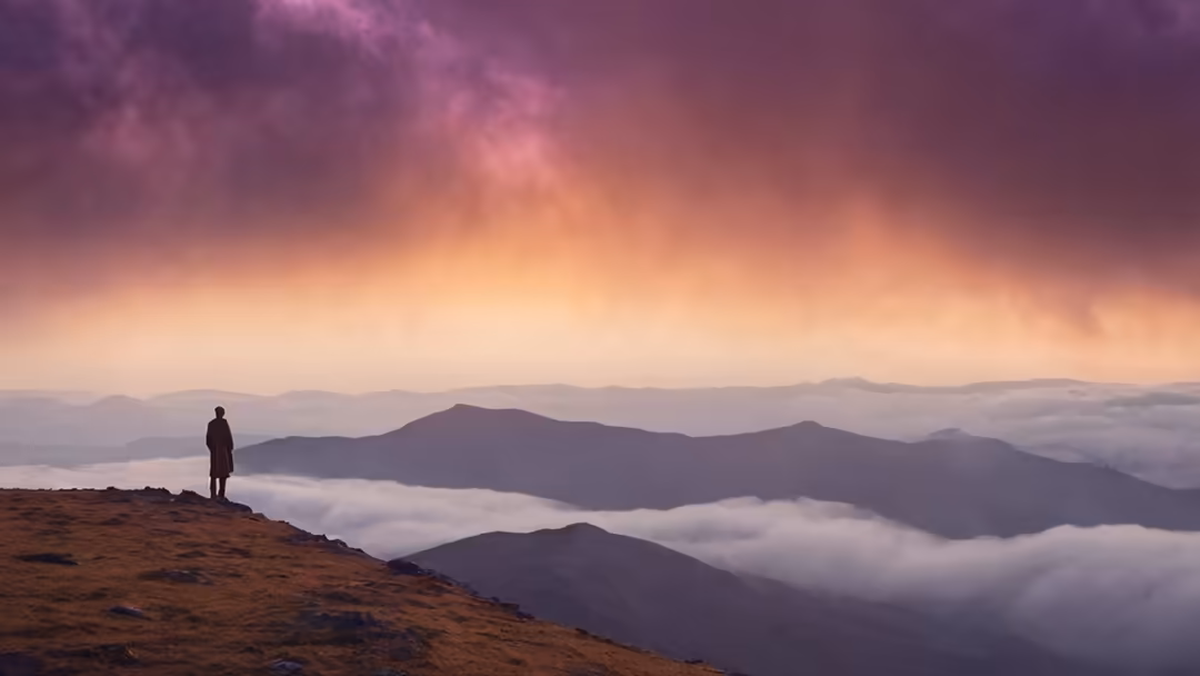 Solitary figure standing on a mountain ridge overlooking misty valleys with dramatic purple and golden sunrise sky above laye