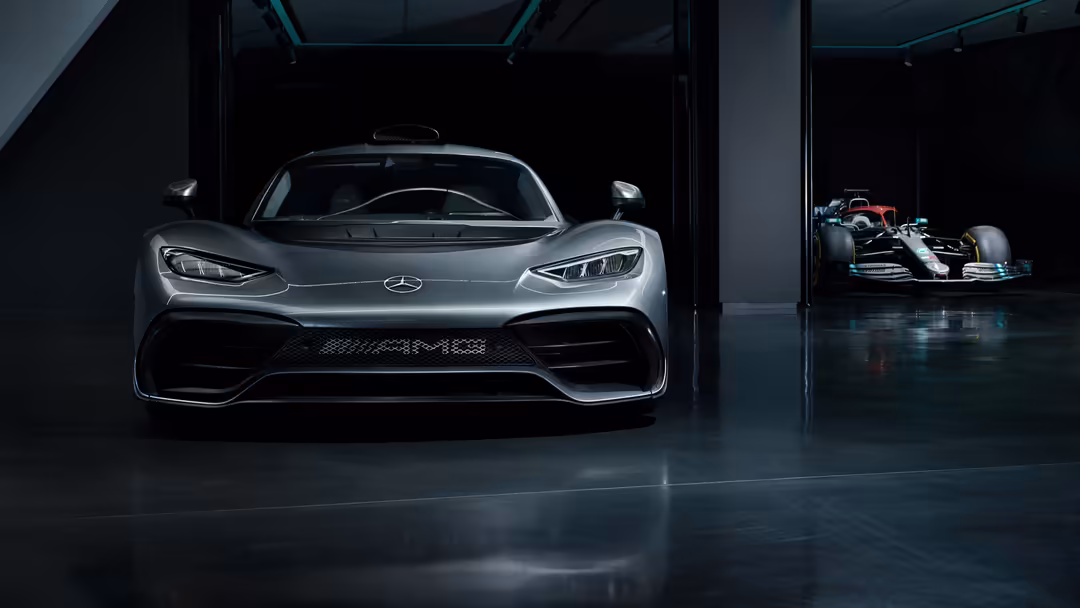 Silver Mercedes-AMG One hypercar parked in modern garage with Formula 1 car visible in background, sleek design and dramatic