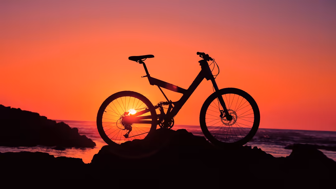 Mountain bike silhouetted against vibrant orange and pink sunset sky over rocky coastal landscape at dusk