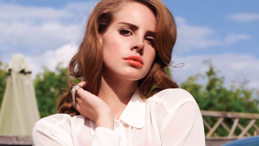 Woman with reddish-brown wavy hair wearing a white collared shirt, gazing pensively against a blue sky and green garden backd