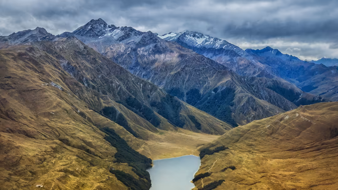 Snow-capped mountain peaks surrounding a turquoise alpine lake nestled in a lush green valley with dramatic cloudy skies over