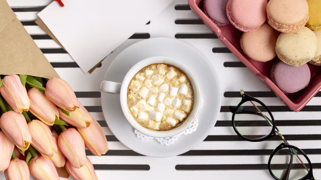 Flat lay arrangement of pastel pink macarons in carton box with hot chocolate, pink tulips, and black-framed glasses on strip