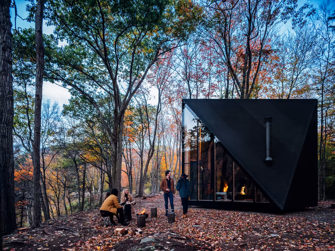 Friends gathering around a campfire outside a modern black cabin nestled in an autumn forest with colorful fall foliage and w