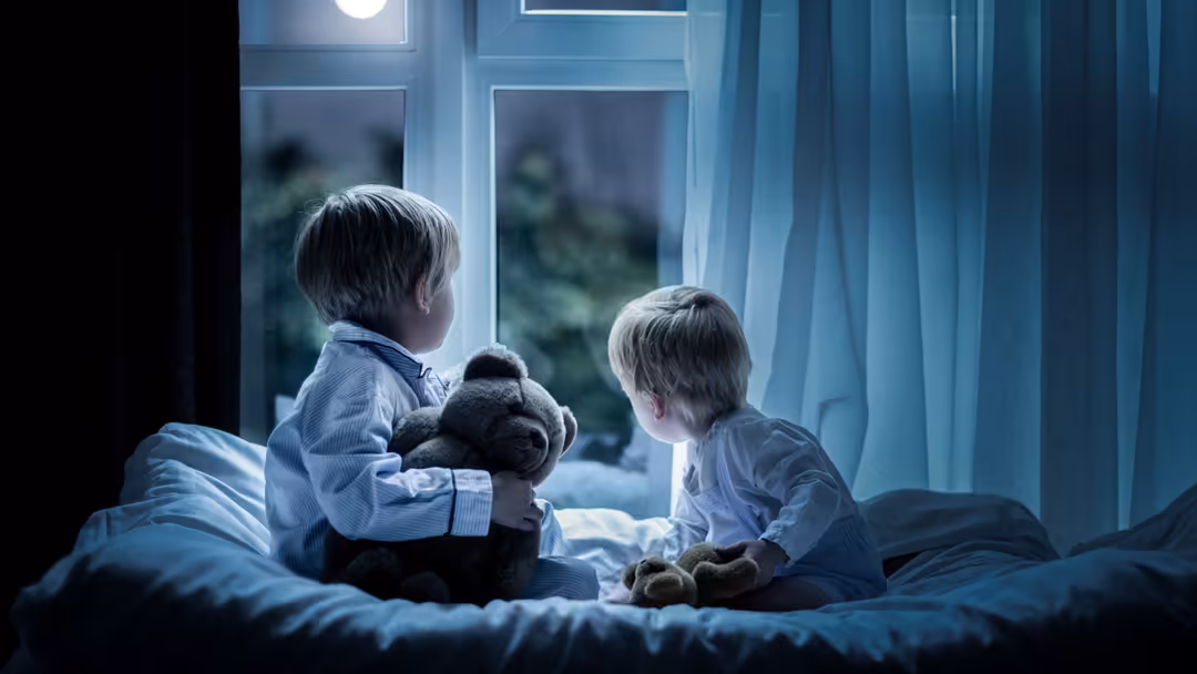 Two young children sitting on a bed at night, holding a stuffed bear, gazing out a moonlit window with sheer blue curtains
