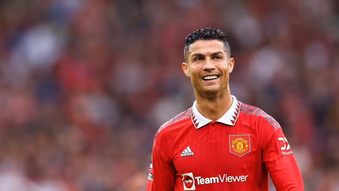 soccer player in red Manchester United jersey with white collar smiles at stadium with blurred crowd behind.