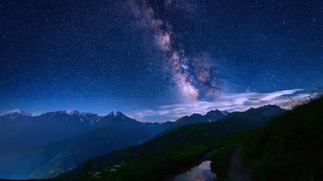 Alpine mountain range with snow-capped peaks under a dramatic starry night sky featuring the luminous Milky Way galaxy arcing
