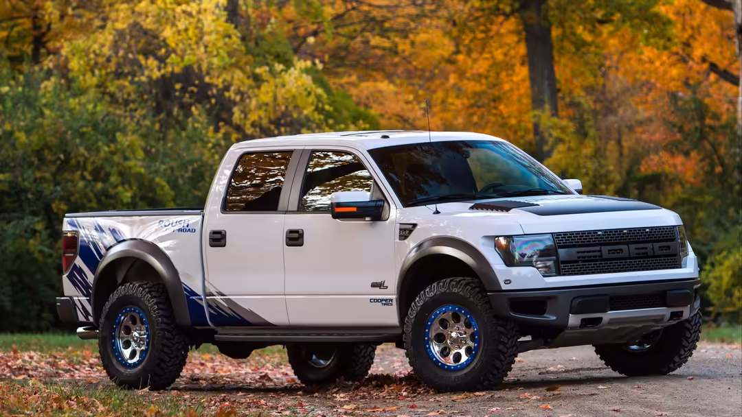 White pickup truck with blue accents parked on autumn road surrounded by trees with golden and orange fall foliage