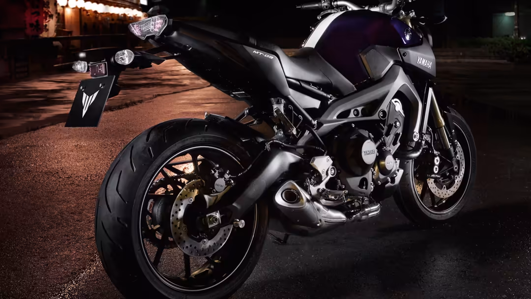 Black motorcycle parked on wet street at night with warm street lights and neon signs in background, sleek modern design
