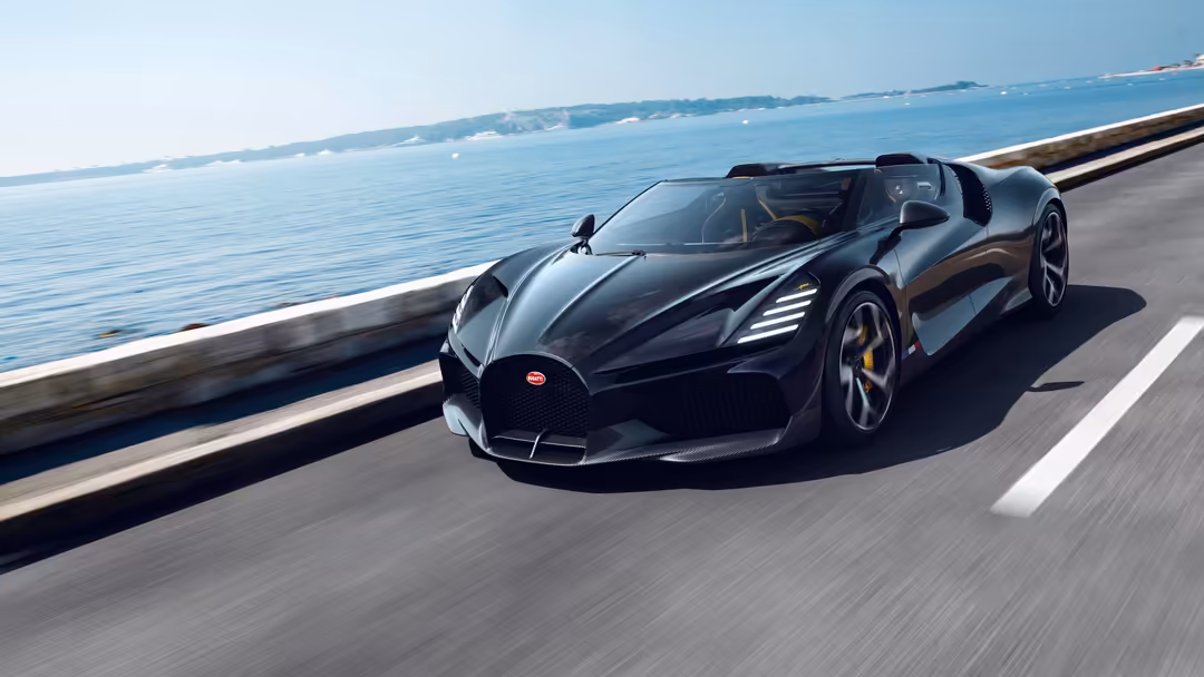 Black Bugatti W16 Mistral convertible driving on coastal highway with ocean and blue sky in background