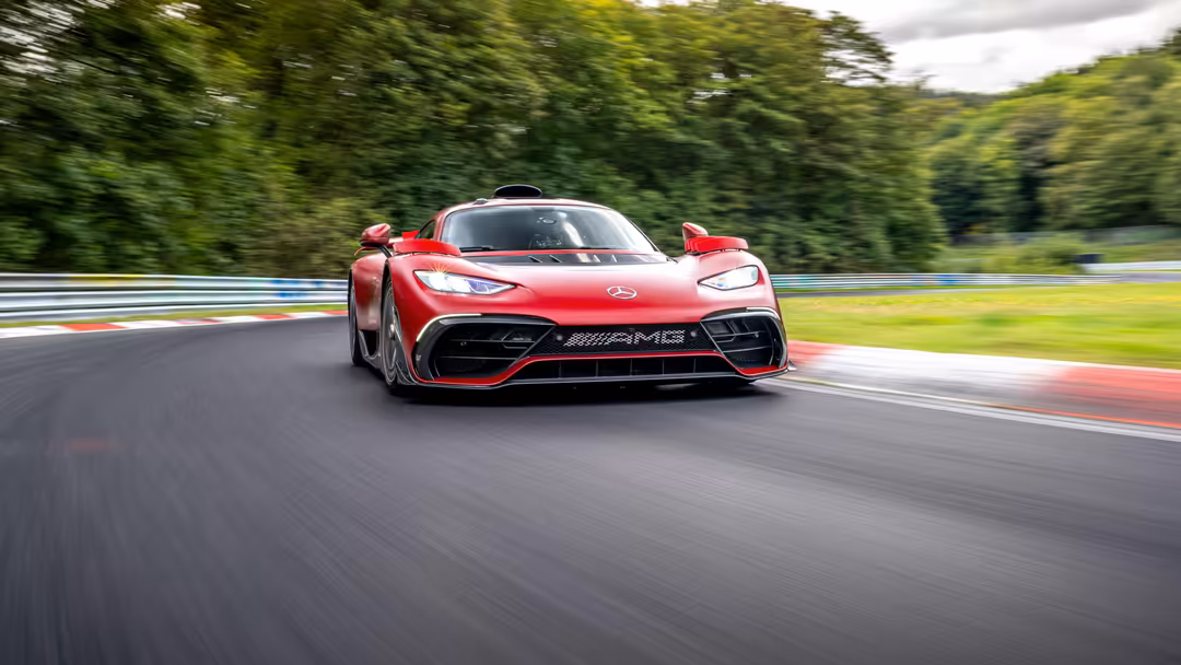 Red Mercedes-AMG One hypercar accelerating on race track with motion blur, surrounded by green trees and cloudy sky