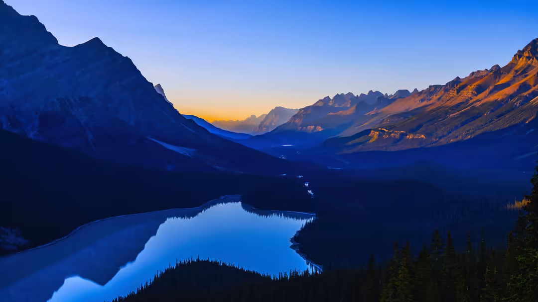 Majestic alpine mountain valley with pristine blue lake reflecting golden sunset light on jagged peaks and forested slopes at